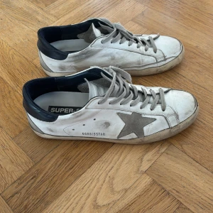 Vita sneakers med grå stjärna från Golden Goose - Golden Goose sneakers i vitt skinn med grå stjärna på sidan och mörkblå häl. Skorna har snörning, rund tå och platt sula. Klassisk design med tydliga Golden Goose-detaljer och medföljande dustbag.