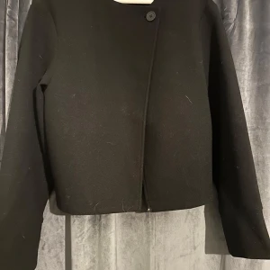 Svart kavajjacka från Gina Tricot - Säljer en svart, croppad kavajjacka från Gina Tricot i storlek XS. Jackan har lång ärm, enkel knäppning med en knapp upptill och rak passform. Perfekt för dig som gillar stilrena och minimalistiska plagg.