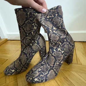 Ormskinnsmönstrade boots med klack - Säljer ett par höga boots med chunky klack i ormskinnsmönster. Skorna har rund tå och är i syntetmaterial. Perfekta för dig som vill sticka ut med ett djurmönster i beige och svart. Aldrig använd från H&M.