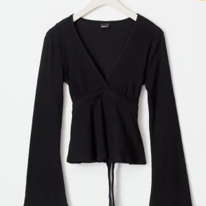 Tie top från Gina tricot - Svart topp med djup v-ringning och långa ärmar. Modellen har en lätt utsvängd passform nedtill och knytband i midjan. Perfekt att styla med jeans eller kjol.