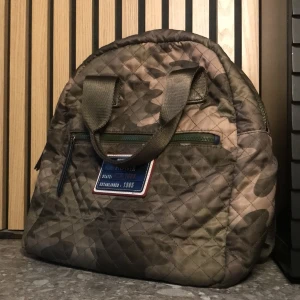 Quiltad ryggsäck i camouflage från Tommy Hilfiger - Säljer en quiltad ryggsäck från Tommy Hilfiger i camouflage-mönster. Väskan har både handtag och axelremmar, dragkedja framtill och en patch med texten 'Hilfiger New York'. Perfekt för dig som vill ha en cool och praktisk accessoar.