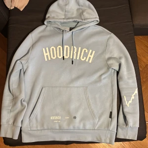 Ljusblå hoodie från Hoodrich - Säljer en ljusblå hoodie från Hoodrich med tryck både fram och bak. Tröjan har huva med dragsko, magficka och ribbade muddar. Stora vita logotyper och textdetaljer på bröstet, ryggen och ena ärmen.