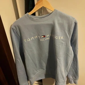 Ljusblå sweatshirt från Tommy Hilfiger - Säljer en ljusblå sweatshirt från Tommy Hilfiger med tryckt logga och text på bröstet. Tröjan har rund halsringning och långa ärmar. Perfekt för en avslappnad och stilren look.