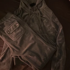 Mjukisbyxor i grå velour med fickdetalj - Grå mjukisbyxor i mjuk velour med en stor ficka med knapp på benet. Byxorna har resår i midjan och en avslappnad passform, perfekta för en chill stil.Juciy sett. Pris 1350kr vid snabb köp kan priset minskas.storlek S