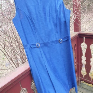 Kornblå ärmlös klänning med bältdetalj - Säljer en kornblå ärmlös klänning med markerad midja och dekorativt bälte med två metallringar framtill. Klänningen har en rak passform och dragkedja i ryggen. Perfekt för dig som gillar stilrena och klassiska plagg.