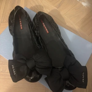 Svarta ballerinaskor från Prada - Snygga svarta ballerinaskor från Prada med en stor rosett framtill. Skorna har en rund tå och är tillverkade i ett glansigt material som ger en elegant touch. Perfekta för att lägga till en stilfull detalj till din outfit.