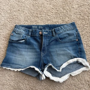 Jeansshorts med spetskant  - Blå jeansshorts från med smal passform och dekorativ vit spetskant längst ner. Klassisk femficksmodell med knapp och dragkedja framtill. Perfekta för varma dagar. Använda men bra skick. Shorts för barn/ungdomar 