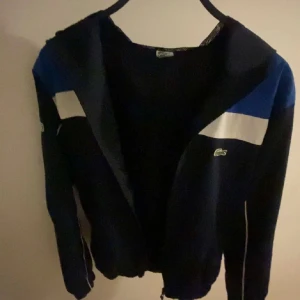 Blå vindjacka från Lacoste - Säljer en blå vindjacka från Lacoste med vit och blå panel upptill samt dragkedja framtill. Jackan har huva och två sidofickor. Klassisk Lacoste-logga på bröstet och sportig design.