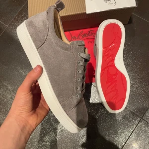  Christian Louboutin skor - Säljer ett par gråa sneakers från Christian Louboutin med klassisk röd sula. Skorna har snörning och är tillverkade i mocka med vita gummisulor. Perfekta för dig som vill ha en stilren och lyxig look.