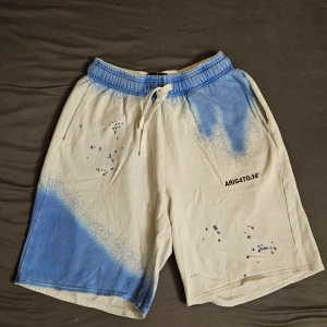 Vita och blå shorts från Axel Arigato - Säljer ett par unika vita shorts från Axel Arigato med blå spray-effekt och trycket 'ARIG4TO:14' på ena benet. Shortsen har resår i midjan med snörning och sidofickor. Perfekta för en avslappnad stil.