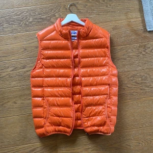 Orange dunväst från Varsity Series - Säljer en orange dunväst från Varsity Series. Västen har en quiltad design med ståkrage och dragkedja framtill. Perfekt för lager-på-lager och ger en sportig look.