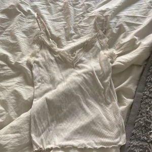 Vit ribbad linnetopp med spets från Amisu - Säljer en söt vit ribbad linnetopp från Amisu med tunna axelband och spetsdetaljer längs urringningen. Perfekt att styla med jeans eller kjol för en fräsch look. Superfin och enkel att matcha!