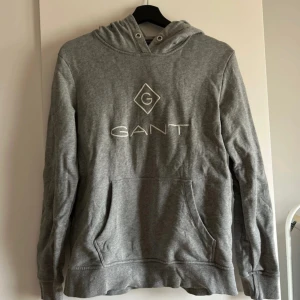 Grå hoodie från GANT - Säljer en klassisk grå hoodie från GANT med broderad logga framtill. Tröjan har huva med dragsko och en stor magficka. Perfekt för en avslappnad stil och enkel att matcha med det mesta.