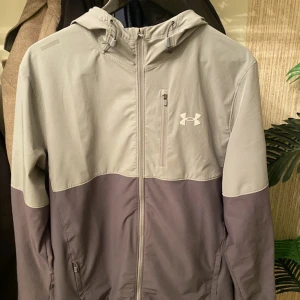 Grå och mörkgrå vindjacka från Under Armour - Snygg tvåfärgad vindjacka från Under Armour i grått och mörkgrå. Jackan har huva, dragkedja framtill och två sidofickor med dragkedja. Perfekt för utomhusaktiviteter och sportiga dagar.