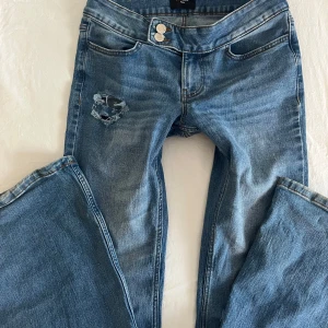 Blå lågmidjade utsvängda jeans med slitningar - Säljer ett par blå jeans med coola slitningar på låret och knät. Dem är lågmidjade och använda någon enstaka gång.
