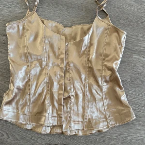 Guld satinlinnetopp från Gina - Säljer en glansig beige linnetopp från Gina i satinmaterial. Toppen har smala axelband och knäppning framtill, perfekt för sommaren. Snygg passform med sömdetaljer som ger en stilren look.