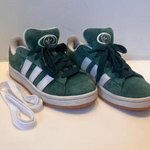 Adidas campus gröna sneakers - Trendiga göna Adidas Campus sneakers med vita ränder och vit sula. Ovandel i mocka och klassisk Adidas-logga på plösen och hälen. Skorna har gröna skosnören och kommer med extra vita skosnören. De vita skosnören är aldrig använda. Dessa var mina favoriter förra sommaren men köpte dem tyvärr för små så de kom inte till så mycket användning😢Därav bra skick! Nypris se på bild (1449kr). Skorna är perfekta för en trendig look och är enkla att styla till allt!🫶🏼