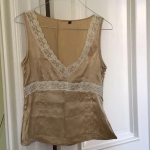 Beige sidenlinne med spetsdetaljer - Elegant beige linne i 100% siden med v-ringning och vackra spetsdetaljer. Perfekt för en stilren look. Linnet är ärmlöst och har en smickrande passform.