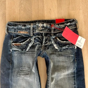 Blå lågmidjade bootcut jeans  - Snygga blå bootcut jeans från El Lago med slitna detaljer och patchar. De har en låg midja och dubbla knappar i gylfen. Perfekta för en avslappnad stil med en touch av edge. Inga bilder på då dom är för små