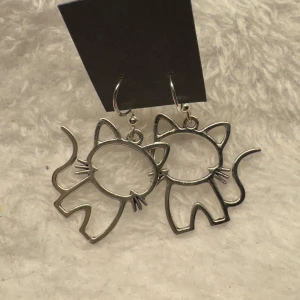 Söta kattörhängen i silverfärg - Ett par örhängen i form av stiliserade katter i silverfärgad metall. Perfekta för dig som älskar katter och vill ha ett lekfullt smycke som sticker ut. Hängande modell med öppna konturer och fina detaljer som morrhår och svans.