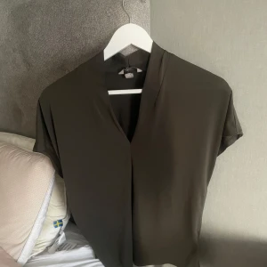 Mörkgrön blus med v-ringning - Säljer en stilren mörkgrön blus med v-ringning och korta ärmar. Blusen har en lös passform och är tillverkad i ett mjukt, lätt material som ger ett snyggt fall. Perfekt att styla till olika outfits!