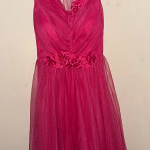 Rosa klänning med blommiga detaljer - Snygg rosa klänning med v-ringning och blommiga detaljer vid midjan. Klänningen har en luftig kjol och är perfekt för festliga tillfällen. Den är ärmlös och har en elegant design som drar blickarna till sig.