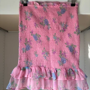 Rosa blommig kjol med volang - Supersöt rosa kjol med blommigt mönster i lila, blått och grönt. Kjolen har smockad midja och dubbla volanger längst ner för extra volym. 