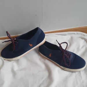 Mörkblå sneakers från Polo Ralph Lauren - Säljer ett par mörkblå sneakers från Polo Ralph Lauren med orange logga på sidan och matchande snörning med orange detaljer. Skorna har rund tå och platt sula, perfekt för en stilren vardagslook.
