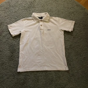 Vit pikétröja från Peak Performance - Klassisk vit pikétröja från Peak Performance med broderad logga på bröstet. Tröjan har krage, knappar framtill och är kortärmad. Perfekt för en sportig och stilren look. Även knappar som går att öppna med. 