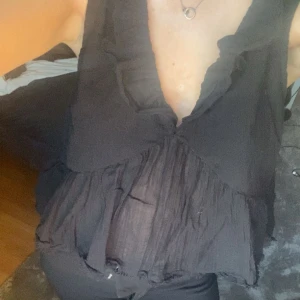 Svart blus med volangdetaljer - Säljer en svart blus med volangdetaljer och djup v-ringning. Blusen har en knytning i ryggen och är ärmlös, vilket ger en luftig och elegant look. Perfekt för en stilren outfit. Den är i storlek L men är precis som s eller xs så jag har dom storlekarna så tycker inte den är stor eller något!!