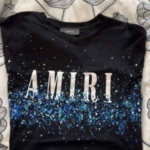 Svart t-shirt från Amiri med blått stänkmönster - Snygg svart t-shirt från Amiri med ett coolt blått stänkmönster och stort vitt tryck på framsidan. Tillverkad i 100% bomull med material från Italien. Perfekt för en stilren och modern look.