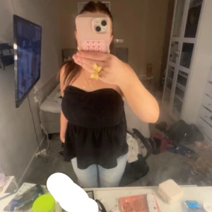 Svart axelbandslös topp - Snygg svart axelbandslös topp med volangdetalj. Perfekt för en stilren look. Toppen har en smickrande passform och är lätt att matcha med olika outfits.