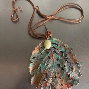 Halsband med lövformat hänge i koppar - Unikt halsband med ett stort lövformat hänge i patinerad koppar och en ljus sten. Hängs på ett brunt läderband som kan justeras i längd. Perfekt för dig som gillar naturinspirerade smycken och boho-stil.