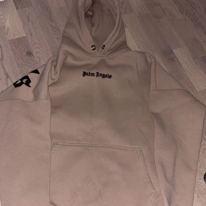 Beige hoodie från Palm Angels - Säljer en beige hoodie från Palm Angels med svart logga på bröstet och stor text på ryggen. Hoodien har magficka och justerbar huva med snören. Perfekt för en avslappnad och trendig stil.