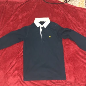 Mörkblå pikétröja från Lyle & Scott - Säljer en mörkblå långärmad pikétröja från Lyle & Scott med vit krage och klassisk gul logga på bröstet. Tröjan har en stilren look och passar perfekt till både jeans och chinos.