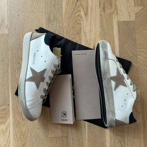 Golden Goose sneakers med beige stjärna - Säljer ett par vita Golden Goose sneakers med beige stjärna på sidan och grå/beige detaljer vid tå och häl. Skorna har snörning och svart hälparti. Klassisk och stilren modell med låg profil.