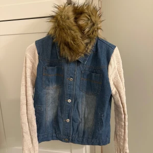 Jeansjacka med stickade ärmar och fuskpäls - Säljer en unik jeansjacka från ELA med ljusbeige stickade ärmar och avtagbar krage i fuskpäls. Jackan har knappar framtill och fyra fickor. Perfekt för dig som vill sticka ut med en cool och annorlunda stil.