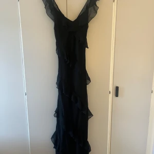 Svart långklänning från Boohoo - Elegant svart långklänning från Boohoo med volangdetaljer och v-ringning. Klänningen har en öppen rygg och är tillverkad i ett lätt, transparent material som ger en luftig känsla. Perfekt för speciella tillfällen som bal