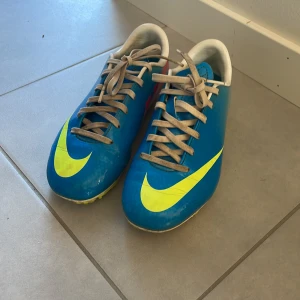 Blå fotbollsskor från Nike - Säljer ett par blå fotbollsskor från Nike med en iögonfallande gul swoosh. Skorna har snörning och är perfekta för fotbollsplanen. De har en rund tå och är designade för att ge bra grepp och komfort under spelet.