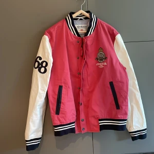 Röd och vit bomberjacka från Morris - Snygg röd och vit bomberjacka från Morris med broderad logga på bröstet och siffran 68 på ärmen. Jackan har långa ärmar och stängs med knappar&dragkedja. Den har ribbade muddar i svart och vitt vid ärmslut och nederkant.