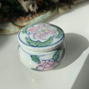Liten porslinsburk med blommönster - Det Charmig liten porslinsburk med ett vackert blommönster i rosa och grönt. Perfekt för att förvara småsaker eller som en dekorativ detalj i hemmet. Tillverkad i Kina.
