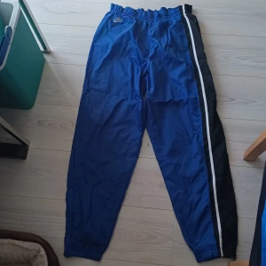 Blå träningsbyxor från Nike - Säljer ett par blå träningsbyxor från Nike med elastisk midja och svarta sidopaneler med vita ränder. Perfekta för sport och fritid.