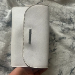 Vit axelväska från Björn Borg - Snygg vit axelväska från Björn Borg med stilren design och silverfärgad logga framtill. Väskan har en rundad lockform och medföljande vit axelrem. Perfekt storlek för det viktigaste och passar till många olika outfits.