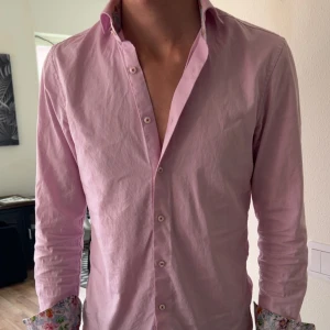 Rosa Skjorta - Säljer en ljusrosa skjorta med blommiga manschetter och insida av kragen. Skjortan har knappar framtill och är långärmad. Perfekt för dig som vill sticka ut lite extra med snygga detaljer. Skjortan är från Stenströms.