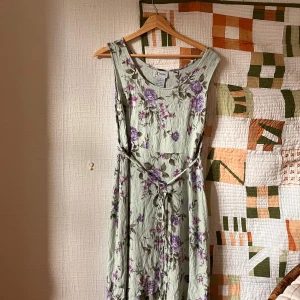 Ljusgrön blommig långklänning K Studio - Superfin långklänning från K Studio i ljusgrönt med lila och rosa blommor. Klänningen är ärmlös, har rund halsringning och ett knytband i midjan. Materialet är 100% rayon med en lätt skrynklig struktur som ger en chill vibe.