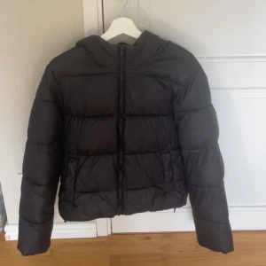 Svart pufferjacka från Gina Tricot - Svart pufferjacka från Gina Tricot i storlek S. Jackan har huva, raka kanter och breda, quiltade sektioner. Stängs med dragkedja framtill och har långa ärmar. Perfekt för kalla dagar när du vill hålla dig varm och ändå se chill ut.