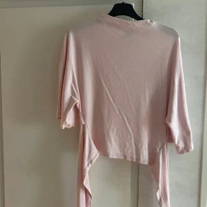 Ljusrosa omlottopp med knytband - Säljer en ljusrosa omlottopp med knytband i midjan och trekvartsärm. Toppen har ett mjuk material. Den är använd 1 gång.