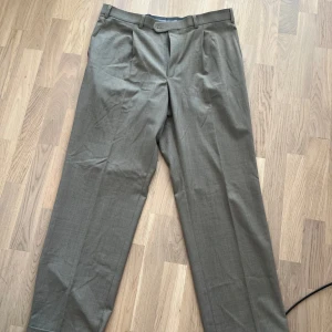 Grå/beige kostymbyxor från Oscar Jacobson - Snygga grå kostymbyxor från Oscar Jacobson i klassisk rak modell. Byxorna har pressveck, bälteshällor och stängs med knapp och dragkedja. Materialet är en mix av ull, polyester och lycra som ger en skön och stilren känsla. Perfekta för dig som gillar dressad stil.