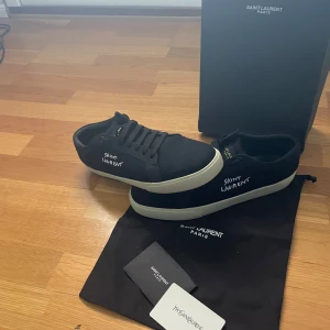 Saint Laurent svarta sneakers strl 44 - Svarta sneakers från Saint Laurent med vit sula och handskriven logga på sidan. Snygg, minimalistisk design med rund tå och snörning. Materialet är canvas och sulan är i gummi. Perfekt för dig som gillar stilrena och exklusiva sneakers med street vibe. Box, dustbag och saint Laurent ättiketer medföljer.