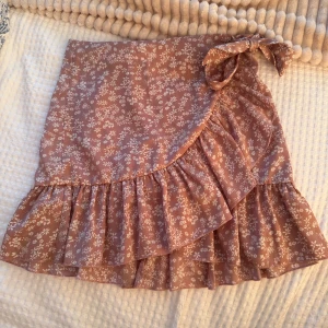 Rosa blommig omlottkjol med volang - Supersöt kort omlottkjol i ljusrosa med vitt blommönster. Kjolen har volangkant och knyts i sidan med en rosett. Perfekt för vår och sommar när du vill ha en romantisk och trendig look.